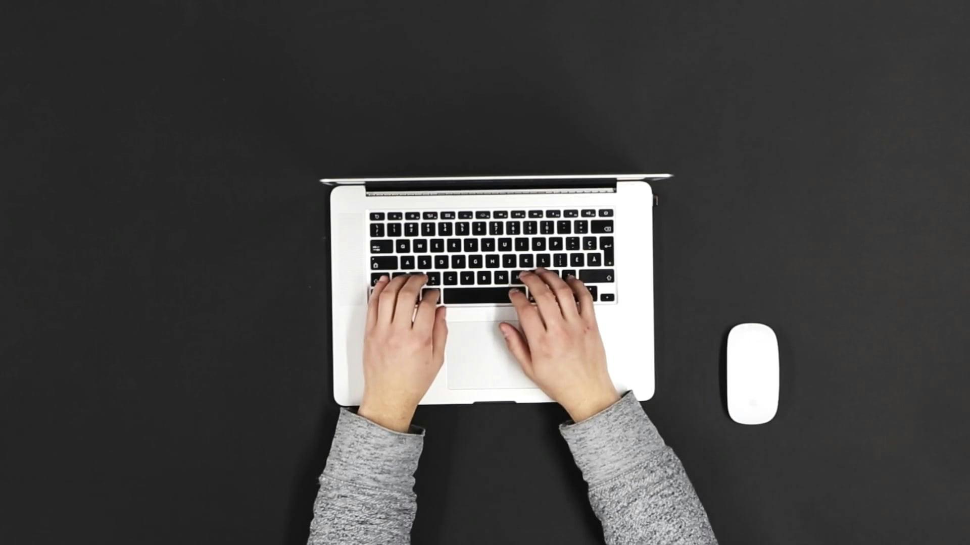 Overhead View Of Hands Typing On A Laptop With Mouse On Dark Surface.